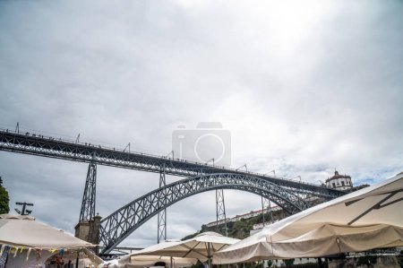 Téléchargez les photos : Toits en toile du marché traditionnel de Ribeira, sur la promenade Vila de Gaia à Porto, avec le pont Dom Luis I en arrière-plan avec des piétons marchant sous un ciel nuageux. - en image libre de droit