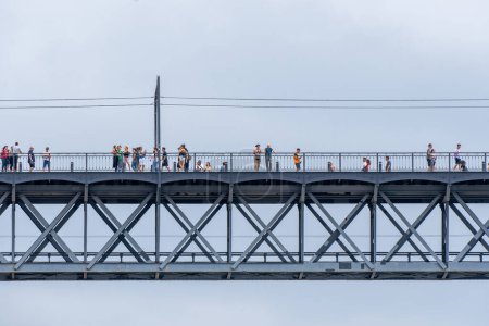 Foto de Turistas caminando, tomando fotos e inclinándose sobre la barandilla del puente de acero Don Luis I en Oporto con nubes de lluvia en el fondo para ver el río Duero. - Imagen libre de derechos