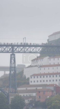 Turistas y peatones caminando con sombrillas y impermeables en la plataforma superior del puente Dom Luis rodeado de niebla y nubes de lluvia baja.