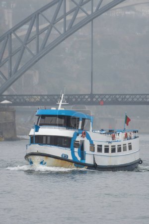Transbordador turístico en primer plano navegando a lo largo del río Duero pasando por debajo del puente de acero Don Luis I en Oporto en un día muy brumoso y lluvioso con tráfico rodando sobre el puente.