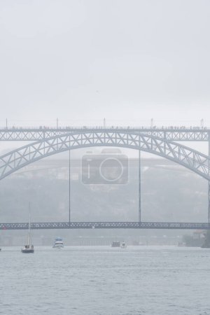 Don Luis I steel bridge wrapped in fog and full of tourists walking with umbrellas and raincoats on a very foggy and rainy day with boats sailing on the Douro River.