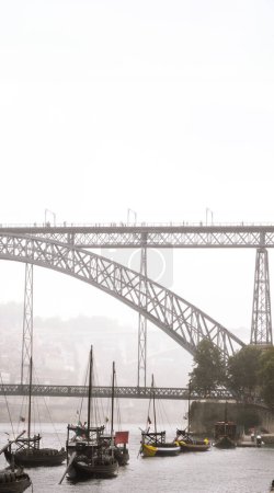 Barcos rabelos de madera atracados en el río Duero de Oporto con barricas de vino en su interior y el puente de acero Don Luis I con turistas paseando con sombrillas envueltas en niebla en un día gris lluvioso.