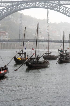 Holzrabelos-Boote mit Weinfässern im Douro-Fluss in Porto, im Hintergrund die Stahlbrücke Don Luis I, die an einem regnerischen grauen Tag in Nebel gehüllt ist.