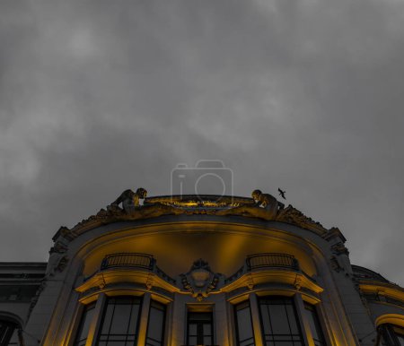 Facade of luxurious classical building, beautifully ornamented with two human sculptures sitting symmetrically on the rooftop illuminated by golden light under a cloudy night sky in the city of Porto.