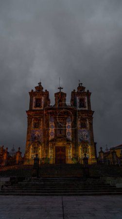 Vista frontal de la Iglesia de San Ildefonso decorada con azulejos portugueses bellamente iluminados con luces cálidas y escalera de granito con puerta de hierro forjado y cruces, bajo un cielo gris y nublado