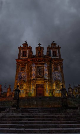 Front view of the Church of San Ildefonso decorated with Portuguese tiles and beautifully illuminated granite staircase with wrought iron fence and crosses, under a gray and cloudy sky. Portugal.