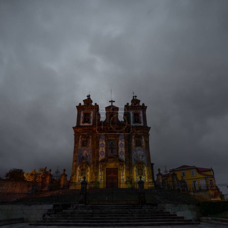 Kirche San Ildefonso und Stufen mit blauen und weißen Keramikfliesen, die in der Dämmerung unter einem grauen wolkenverhangenen Himmel von orangefarbenen Lichtern wunderbar und schwach beleuchtet werden. Portugal.