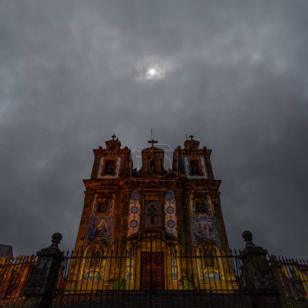 Church of San Ildefonso of Porto covered in blue and white ceramic tiles illuminated beautifully at night, under a cloudy sky with the moonlight illuminating the wrought iron railings. Portugal.