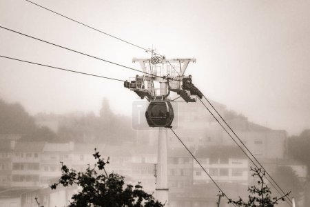 Photo for A Vilanova de Gaia cable car gondola suspended on hanging steel cables ascending under a cloudy sky with the classic Gaia neighborhood shrouded in fog. Black and white. - Royalty Free Image