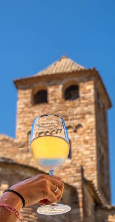 Young woman's hand with ring and bracelets holding and raising a glass of fresh white wine enjoying a sunny day with clear blue sky with a rural village baroque church in the background.