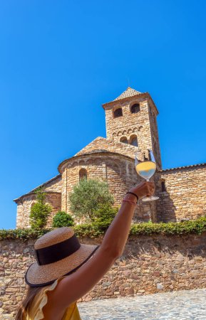Beautiful young woman with long blonde hair wearing a straw hat, raising and toasting with a glass of fresh white wine enjoying a sunny day with a rural village baroque church in the background.