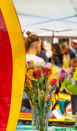 Bouquet of roses decorated with ears of wheat and bows of the flag of Catalonia, at a flower and book stall in a traditional holiday market with people walking on Sant Jordi day.
