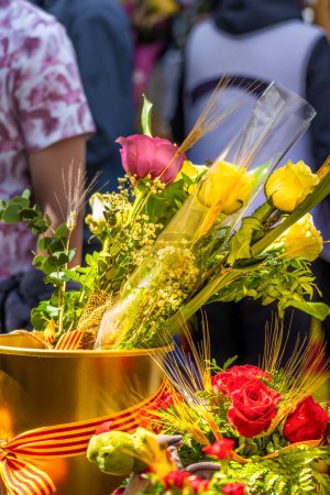 Yellow and red roses in a golden pot with a bow of the flag of Catalonia at a flower stand of the traditional festival of Catalonia with many people walking around on Sant Jordi's day.