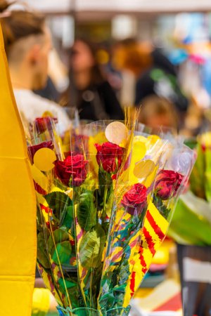 Detail eines Rosenstraußes, geschmückt mit Ähren und der Flagge Kataloniens, in einem Blumen- und Bücherstand auf einem traditionellen Festmarkt mit Menschen, die am Sant Jordi 's Day spazieren gehen.