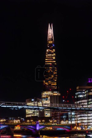 Foto de Vista nocturna del rascacielos de Londres totalmente iluminado The Shard, edificios de oficinas y dos puentes y un ferry sobre el río Támesis con turistas caminando por la noche en Londres. - Imagen libre de derechos