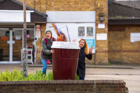 Photo for A middle-aged lesbian couple, joking and teasing, clutching a giant takeaway coffee cup, looking at the camera, smiling and waving outside a London neighborhood supermarket. - Royalty Free Image