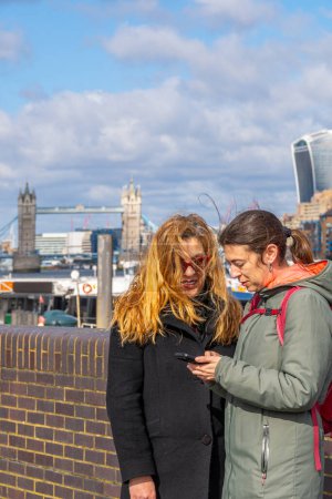 Téléchargez les photos : Un couple de lesbiennes regardant sur leur téléphone portable pour trouver un itinéraire vers le centre de Londres, de la promenade de la rivière Thames avec Tower Bridge en arrière-plan et beaucoup de vent frottant leurs cheveux. - en image libre de droit