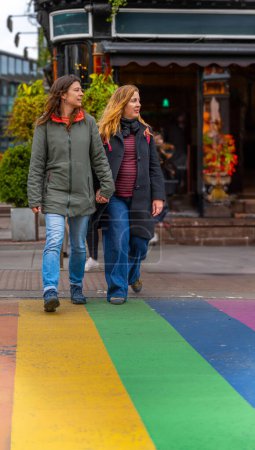 Photo for A happy lesbian couple holding hands, walking and crossing a pedestrian crossing painted with the colors of the rainbow, gay pride and lgbt+ through the Camden Town neighborhood in London. - Royalty Free Image