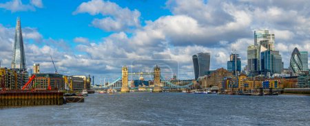 Wide angle panoramic view from the River Thames of the London skyline with skyscrapers and Tower Bridge illuminated by the morning sun and boats and ferries moored at their docks.