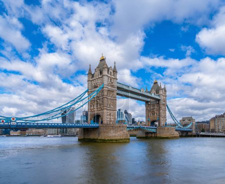 Panoramic view of London's Tower Bridge from the River Thames with maritime traffic and ferries sailing along the river under a blue sky with white clouds. United Kingdom.
