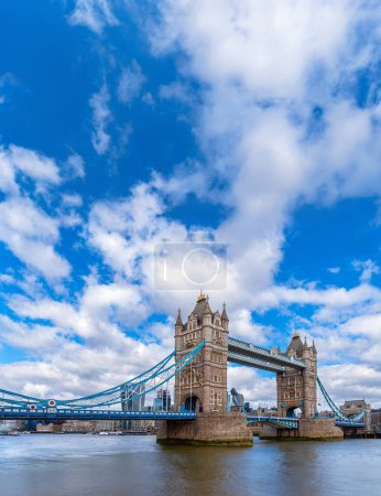Panoramic diagonal view of London's Tower Bridge from the River Thames with maritime traffic and ferries sailing along the river under a blue sky with white clouds. United Kingdom.