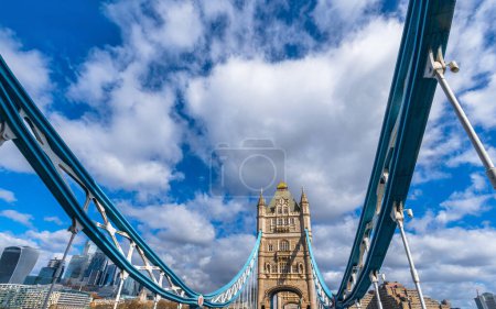 Vista desde abajo y en la calle mirando hacia arriba del Tower Bridge en Londres, con la vista de las estructuras y cables de tensión en perspectiva con la torre iluminada por el sol bajo un cielo azul.