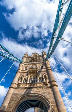 View from below in perspective of the Tower Bridge and the entrance arch of London illuminated by the sun and with the shadows of the steel cables on its facade on a sunny day with white clouds.