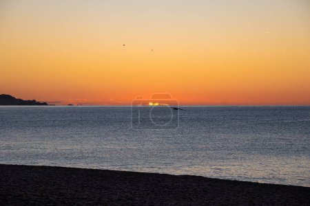Silhouette of seagulls flying across the Mediterranean Sea in calm waters illuminated by the first lights of dawn, creating optical illusions on the sea horizon line under an orange sky.