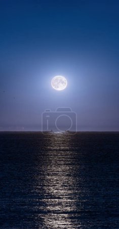 Super full moon in a clear night sky beautifully illuminating the water of the Mediterranean sea and two silhouettes of fishing boats illuminated by the backlit moonlight on the horizon line.