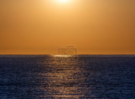 First lights of dawn backlit and illuminating the silhouette of a fishing boat sailing through the calm blue waters of the Mediterranean Sea with the brightness of the sun reflected in its waters.