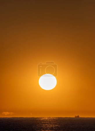 Orangefarbener Himmel mit leuchtend gelber Morgensonne, die ein Fischerboot, das durch das Mittelmeer segelt, erhellt und die ersten Strahlen der Morgensonne, die optische Täuschungen und Fata Morgana am Horizont erzeugen.