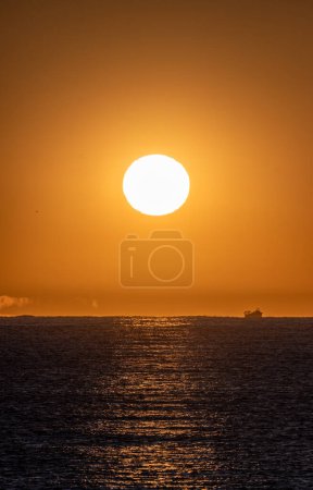 Calm sea illuminated by the orange light of the bright dawn sun and the silhouette of a fishing boat sailing through the Mediterranean Sea and various optical illusions and mirages on the horizon.