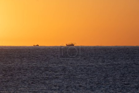 Silhouetten von zwei Fischerbooten, die von der Morgensonne am Horizont des Mittelmeeres beleuchtet werden, um unter einem orangen Himmel nach Garnelen zu fischen.