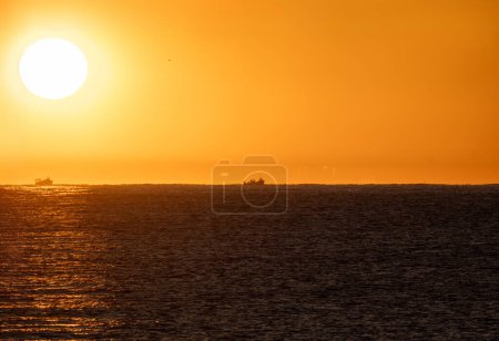 Sol amarillo del amanecer que emerge del horizonte retro del mar Mediterráneo iluminando dos barcos de pesca en silueta que salen a capturar camarones, bajo un cielo naranja.