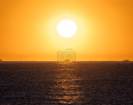 Bright yellow dawn sun rising from the horizon of the Mediterranean Sea illuminating a fishing boats sailing through the calm sunlit water for shrimp fishing, under an orange sky.