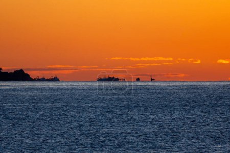 Silhouetten von Fischerbooten, die darauf warten, in See zu stechen, von der Morgensonne an der Horizontlinie des Mittelmeeres beleuchtet, erzeugen Trugbilder und optische Täuschungen unter einem orangefarbenen Himmel.