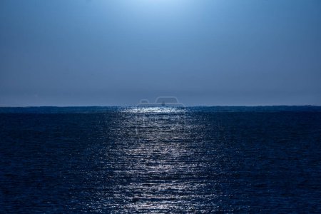 Photo for Clear, bluish night sky illuminated by moonlight with its rays of light beautifully reflected in the water of the Mediterranean Sea and a backlit fishing boat silhouette on the horizon line. - Royalty Free Image