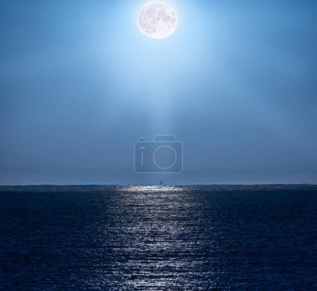 Full moon illuminating the clear, bluish night sky and with its rays of light beautifully reflected in the water of the Mediterranean Sea and illuminating a backlit fishing boat on the horizon line.