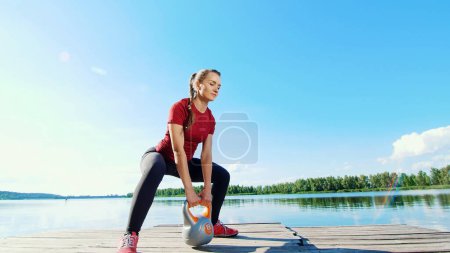 Photo for Beautiful, athletic young blond woman doing different exercises with weights, dumbbells, lunges, squats. Lake, river, blue sky and forest in the background, summer sunny day. High quality photo - Royalty Free Image