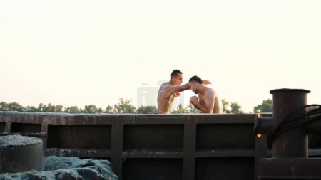 Photo for Young athletic men with bare, naked torsos, box, practice the technique of strikes, capture, fight, on a sandy beach, in a cargo port, near large cargo cranes, at sunrise, in summer. High quality - Royalty Free Image