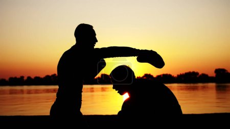 Photo for Two dark male figures, at sunrise, against the light, boxing, fighting in sparring, training in a pair of techniques of strikes. On sandy beach, in cargo port, near water, in summer. High quality - Royalty Free Image