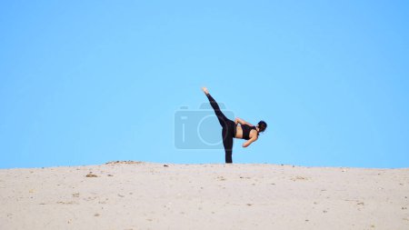 Photo for Athletic girl in a black top and leggings, practice kicks, kicks feet, fight with the shadow, training to fight. On a deserted beach, against a blue sky, in the summer, under a hot sun. High quality - Royalty Free Image