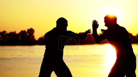 Photo for Two dark male figures, at sunrise, against the light, boxing, fighting in sparring, training in a pair of techniques of strikes. On sandy beach, in cargo port, near water, in summer. High quality - Royalty Free Image