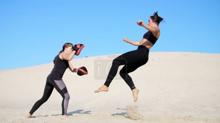 Photo for Two athletic, young women in black fitness suits are engaged in a pair, fulfill kicks, train to fight, on deserted beach, against a blue sky, in summer, under a hot sun. Slow motion. High quality - Royalty Free Image