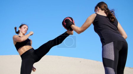 Photo for Two athletic, young women in black fitness suits are engaged in a pair, work out kicks, train to fight, on deserted beach, against a blue sky, in summer, under a hot sun. Slow motion. High quality - Royalty Free Image