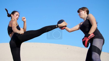 Photo for Two athletic, young women in black fitness suits are engaged in a pair, work out kicks, train to fight, on deserted beach, against a blue sky, in summer, under a hot sun. Slow motion. High quality - Royalty Free Image