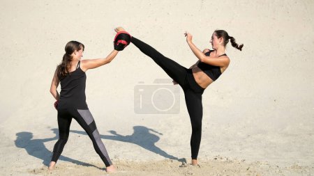 Photo for Two athletic, young women in black fitness suits are engaged in a pair, work out kicks, on a deserted beach, against a blue sky, in the summer, under a hot sun. Slow motion. High quality photo - Royalty Free Image