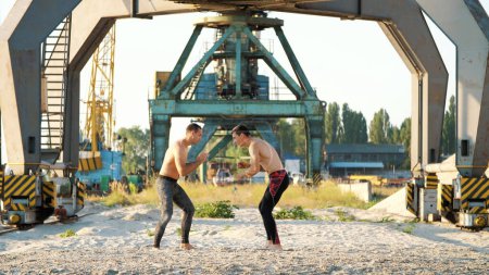 Photo for Young athletic men with bare, naked torsos, box, practice the technique of strikes, capture, fight, on a sandy beach, in a cargo port, near large cargo cranes, at sunrise, in summer. High quality - Royalty Free Image