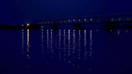 Téléchargez les photos : Nuit, dans l'eau, le pont et ses lumières sont réfléchies. Un long pont sur la rivière. Photo de haute qualité - en image libre de droit