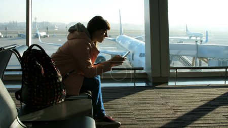 Téléchargez les photos : À l'aéroport, dans la salle d'attente, sur fond de fenêtre donnant sur les avions et la piste, se trouve une jeune femme tapant au téléphone. voir sa silhouette. Photo de haute qualité - en image libre de droit